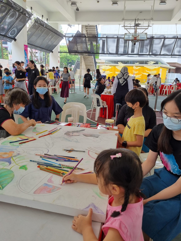 Children and adults collaborate on a large coloring activity at a busy indoor event space with tables, chairs, and art materials